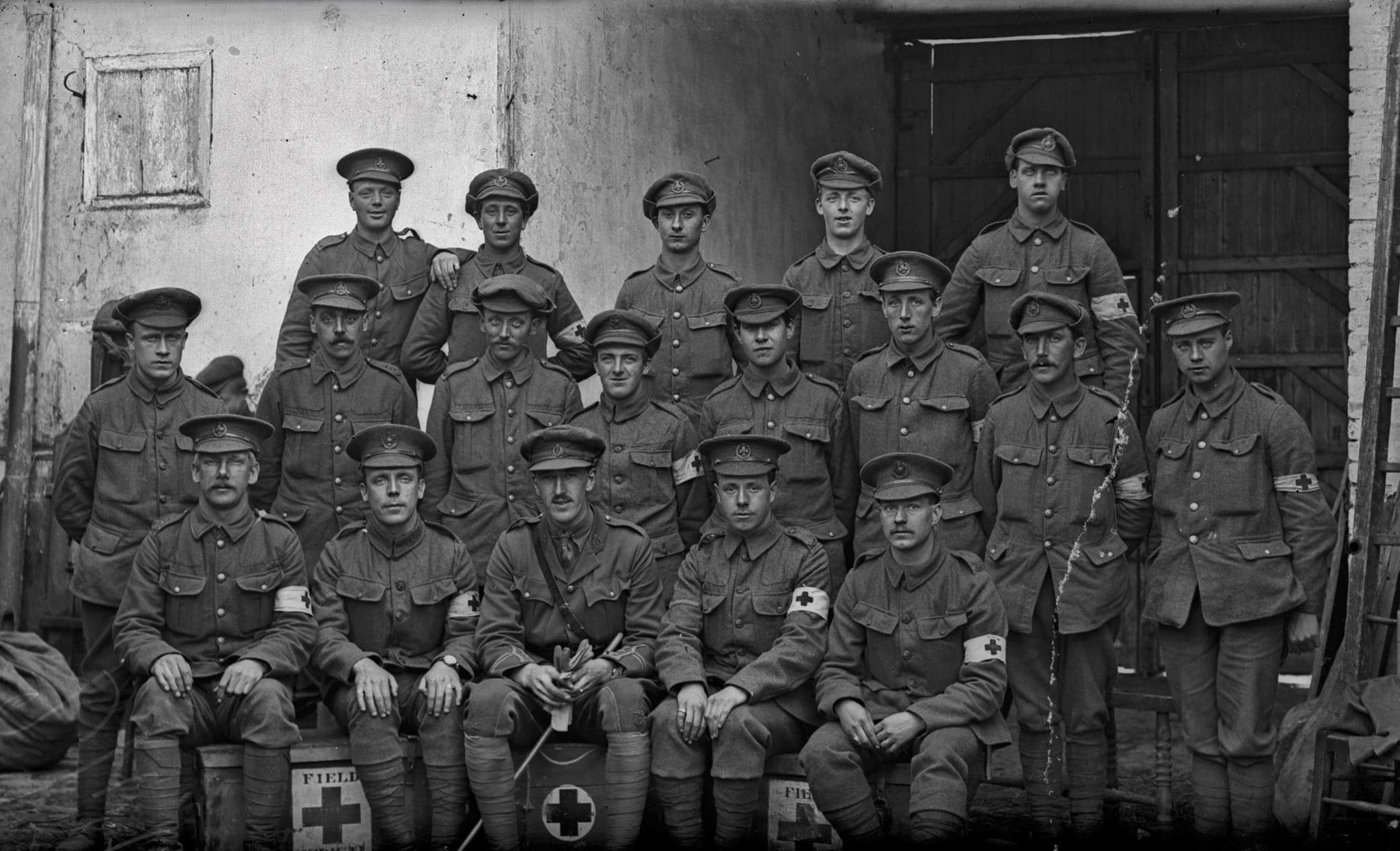 Black and white group photograph of around twenty Leeds Rifles soldiers in First World War service dress and peaked caps, several wearing Red Cross armbands, posed beside a barn with Red Cross field boxes in front of them